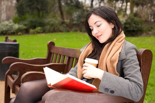 young woman sitting in the park reading a book and drinking a coffee