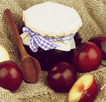 Jar of Plum Jam with Wooden Spoon, Ripe Purple Plums Full Body and Halves closeup on Wicker Sackcloth background