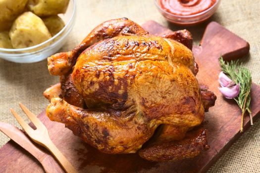 Roast chicken on wooden board with potatoes and ketchup, photographed with natural light (Selective Focus, Focus on the front of the chicken)  