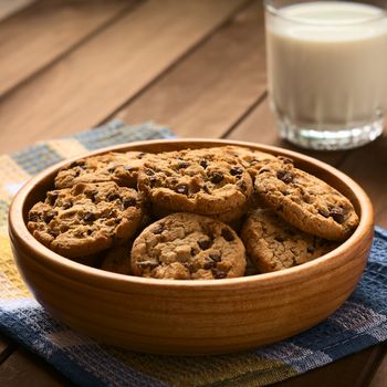 Chocolate chip cookies in wooden bowl with a glass of cold milk in the back, photographed on cloth on wood with natural light (Selective Focus, Focus in the middle of the cookies)
