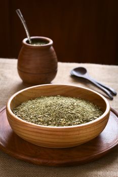 South American yerba mate (mate tea) dried leaves in wooden bowl with a wooden mate cup and strainer (bombilla) in the back, photographed with natural light. Mate is the national infusion of Argentina. (Selective Focus, Focus one third into the dried tea) 