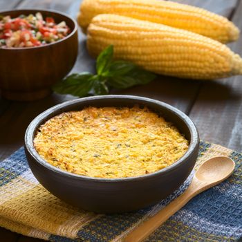 Bowl of traditional Chilean corn pie called Pastel de Choclo served with Pebre sauce in the back, photographed on cloth on dark wood  with natural light. Below the corn-basil mix is ground meat, olive, boiled egg, raisins and pieces of chicken. (Selective Focus, Focus one third into the dish) 