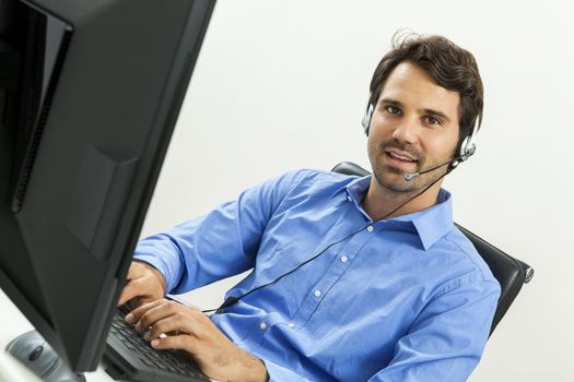 Attractive unshaven young man wearing a headset offering online chat and support on a client services of help desk as he types in information on his computer