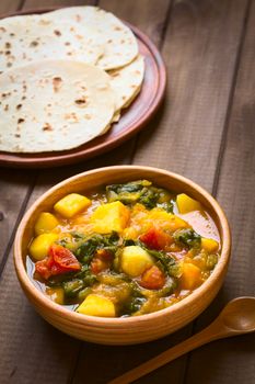 Wooden bowl of pumpkin, mangold, potato and tomato curry dish with homemade chapati flatbread in the back photographed with natural light (Selective Focus, Focus in the middle of the curry dish) 