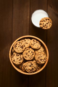 Overhead shot of chocolate chip cookies in wooden bowl and a cookie on the rim of a glass of cold milk, photographed on wood with natural light