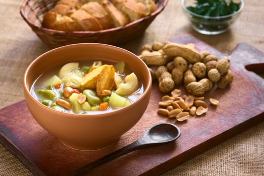 Bowl of traditional Bolivian Sopa de Mani (peanut soup) made of meat, pasta, vegetables (pea, carrot, potato, broad bean, pepper, corn) and ground peanut, photographed on wooden board with natural light (Selective Focus, Focus in the middle of the soup)
