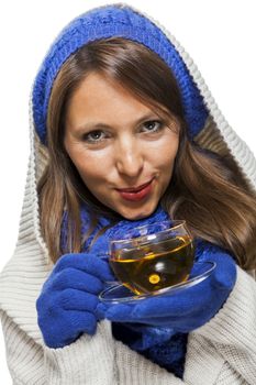 Fashionable young woman in a blue knitted winter ensemble and cowl neck jersey sipping a cup of hot tea with a smile in an effort to keep warm, isolated on white