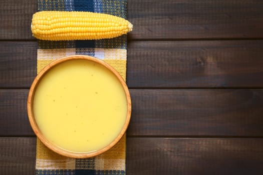 Overhead shot of cream of corn soup in wooden bowl with corn cob on towel, photographed on dark wood with natural light