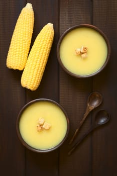 Overhead shot of two bowls of cream of corn soup with croutons on top, wooden spoons and corn cobs on the side, photographed on dark wood with natural light