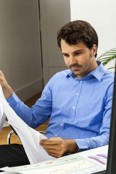 Young handsome man sitting at his desk in the office while reading written agreements and studying important documentation for work