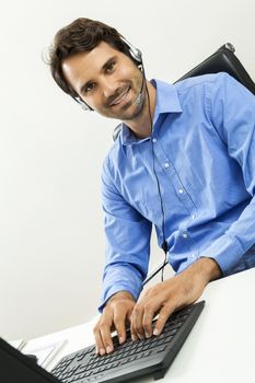 Attractive unshaven young man wearing a headset offering online chat and support on a client services of help desk as he types in information on his computer