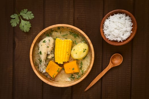 Overhead shot of traditional Chilean Cazuela de Pollo (or Cazuela de Ave) soup made of chicken, sweetcorn, pumpkin and potato, seasoned with fresh coriander served in wooden bowl and rice on the side (which is usually served with the soup), photographed on dark wood with natural light