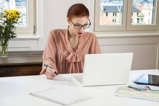 Sitting Young Businesswoman Writing on a Paper While Looking at her Laptop Computer Seriously.