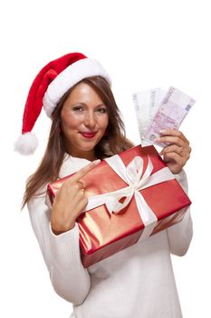 Vivacious beautiful young woman wearing a Santa hat with a large red Christmas gift and money, conceptual of successful Xmas shopping and payment, on white