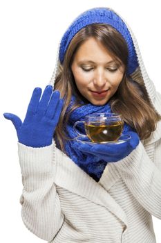 Fashionable young woman in a blue knitted winter ensemble and cowl neck jersey sipping a cup of hot tea with a smile in an effort to keep warm, isolated on white