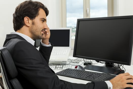 Stylish businessman in a suit sitting at his desk in the office chatting on the phone with a view of his blank computer monitor