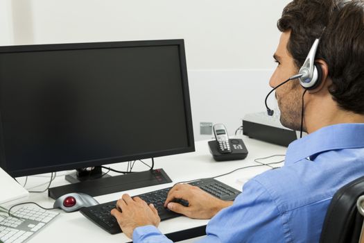 Attractive unshaven young man wearing a headset offering online chat and support on a client services of help desk as he types in information on his computer