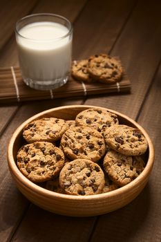 Chocolate chip cookies in wooden bowl with a glass of cold milk in the back, photographed on wood with natural light (Selective Focus, Focus on the cookie in the middle)
