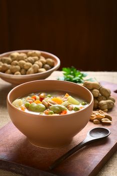 Bowl of traditional Bolivian Sopa de Mani (peanut soup) made of meat, pasta, vegetables (pea, carrot, potato, broad bean, pepper, corn) and ground peanut, photographed on wooden board with natural light (Selective Focus, Focus one third into the soup)