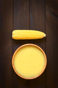 Overhead shot of cream of corn soup in wooden bowl with corn cob, photographed on dark wood with natural light