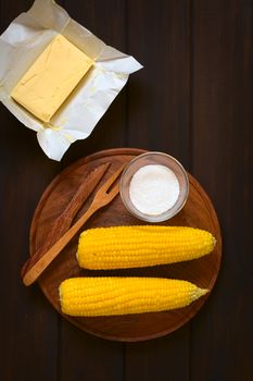 Overhead shot of two cobs of cooked sweet corn on wooden plate with salt and cutlery, with a piece of butter above, photographed on dark wood with natural light