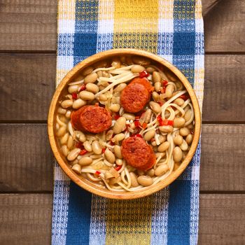 Overhead shot of traditional Chilean dish called Porotos con Riendas (English: beans with reins), made of cooked beans, linguine (flat spaghetti) and served with fried sausage, photographed on wood with natural light