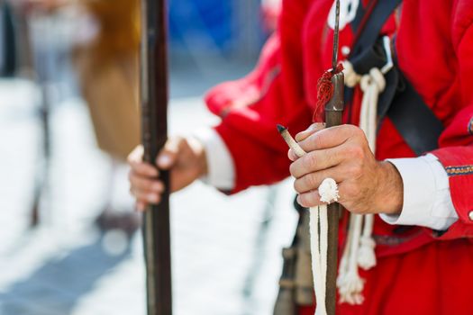 Hands of a musketeer holding a musekt and slow match or match cord with a glowing tip, ready to ignite the gunpowder and fire