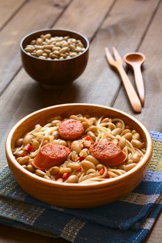 Traditional Chilean dish called Porotos con Riendas (English: beans with reins), made of cooked beans, linguine (flat spaghetti) and served with fried sausage, photographed on dark wood with natural light (Selective Focus, Focus on the two sausage pieces in the front)