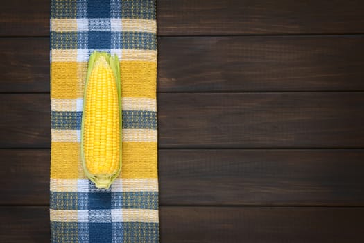 Overhead shot of husked cob of sweet corn photographed on dish towel on dark wood with natural light