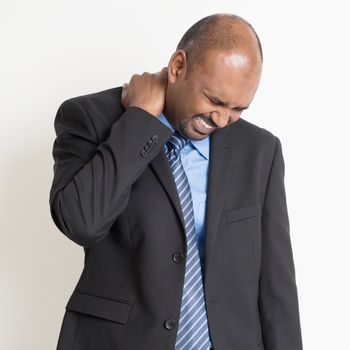 Indian businessman shoulder pain, holding his neck with painful face expression, standing on plain background.