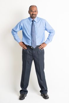 Full body Indian businessman hand on waist looking at camera, standing on plain background with shadow