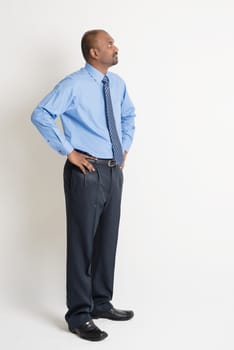 Full body Indian businessman hand on waist looking up at blank copy space, standing on plain background with shadow.