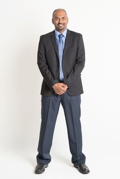 Full length friendly Indian businessman in formal suit looking at camera, standing on plain background.