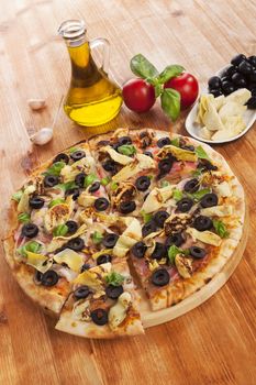 Delicious pizza background. Fresh pizza, garlic, olive oil, fresh herbs, tomatoes, artichokes and black olives on wooden background. 