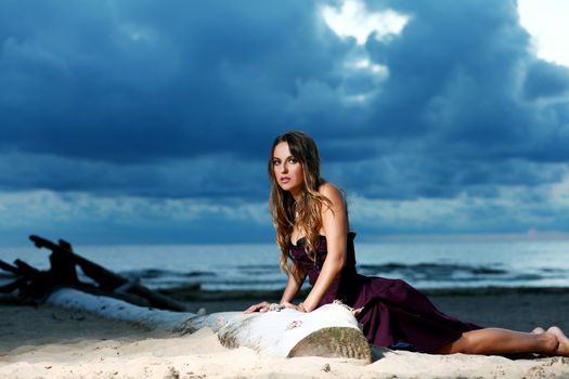 Beautiful girl with brown hair who is wearing a dark purple dress is posing at a beach over a dark sky background