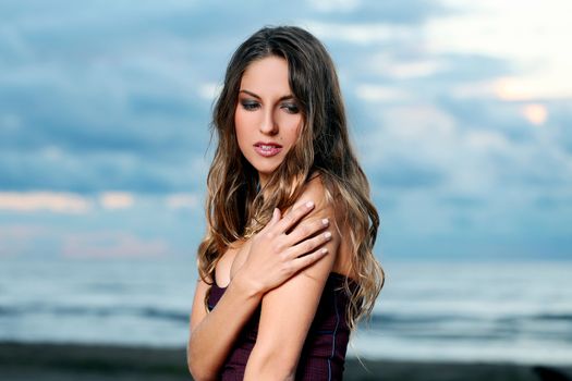 Beautiful girl with brown hair who is wearing a dark purple dress is posing at a beach over a sky background