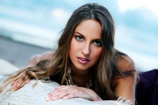 Beautiful girl with brown hair who is wearing a dark purple dress is posing at a beach