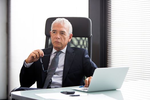 Half length portrait of a thoughtful businessman in the office with laptop computer and holding glasses 