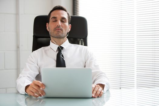 Half length portrait of a male businessman with eyes closed, earphones and laptop computer