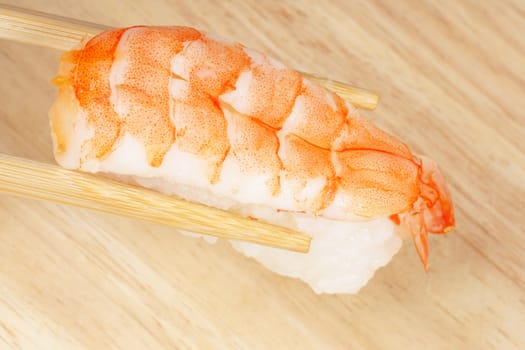 Wooden chopsticks holding a piece of sushi with shrimp, over a wooden background