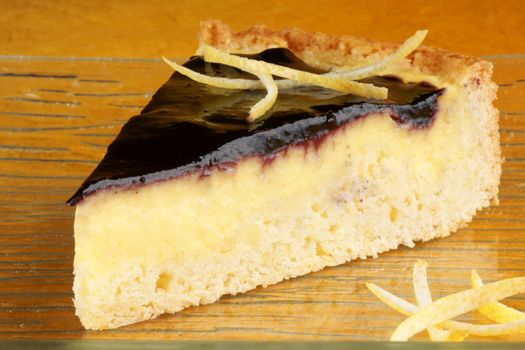Slice of blueberry marmalade and custard cream tart served on a glass plate with lemon zest, over an orange background.