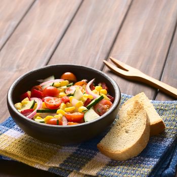 Bowl of fresh vegetable salad made of sweet corn, cherry tomato, cucumber, red onion, red pepper, chives with toasted bread on the side, photographed on dark wood with natural light (Selective Focus, Focus one third into the salad)