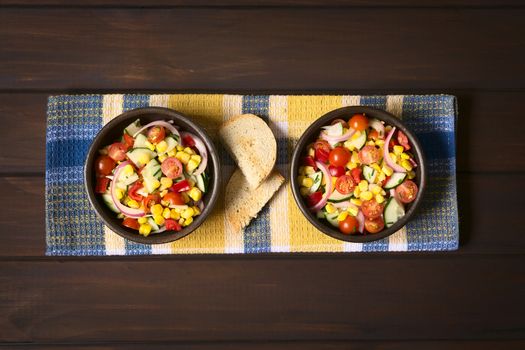 Overhead shot of two bowls of fresh vegetable salad made of sweet corn, cherry tomato, cucumber, red onion, red pepper, chives with toasted bread on the side, photographed on dark wood with natural light