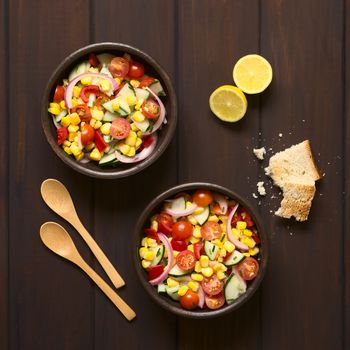 Overhead shot of two bowls of fresh vegetable salad made of sweet corn, cherry tomato, cucumber, red onion, red pepper, chives with toasted bread on the side, photographed on dark wood with natural light