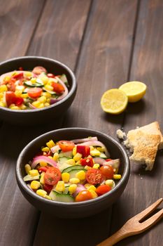 Bowls of fresh vegetable salad made of sweet corn, cherry tomato, cucumber, red onion, red pepper, chives with toasted bread on the side, photographed on dark wood with natural light (Selective Focus, Focus one third into the first bowl)