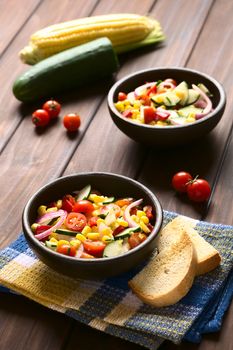 Bowls of fresh vegetable salad made of sweet corn, cherry tomato, cucumber, red onion, red pepper, chives with toasted bread on the side, photographed on dark wood with natural light (Selective Focus, Focus in the middle of the first bowl)