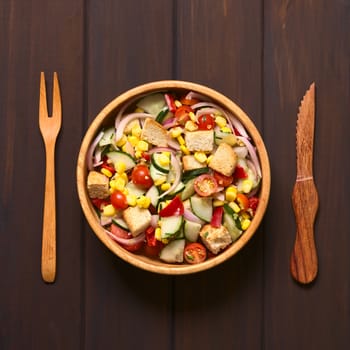 Overhead shot of fresh vegetable salad made of sweet corn, cherry tomato, cucumber, red onion, red pepper, chives with croutons in wooden bowl, photographed on dark wood with natural light