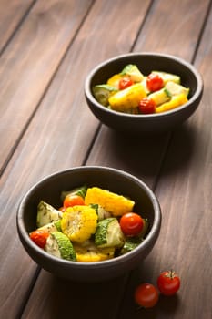 Two bowls of baked vegetables of sweet corn, zucchini and cherry tomato, seasoned with thyme, photographed on dark wood with natural light (Selective Focus, Focus in the middle of the first bowl)