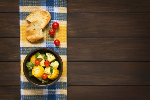 Overhead shot of a bowl of baked vegetables of sweet corn, zucchini, cherry tomato with thyme, toasted bread slices above, photographed on dish towel on dark wood with natural light