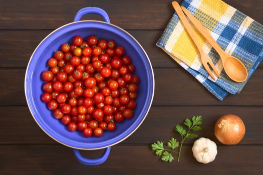 Overhead shot of cherry tomatoes in blue metal strainer with garlic, onion, parsley leaves and wooden cutlery on the side, photographed on dark wood with natural light